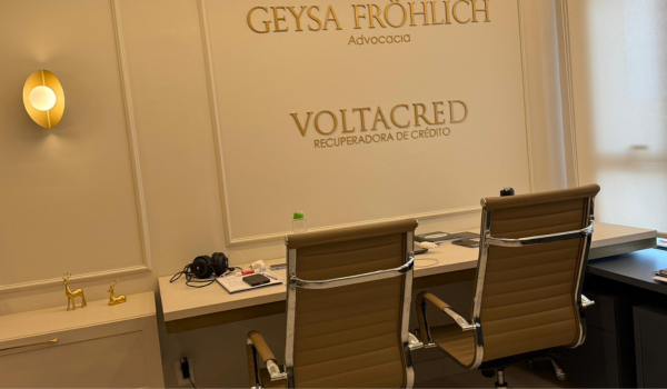 Recepção da sede: parede com as marcas Geysa Fröhlich Advocacia e Voltacred, bancada de atendimento e duas cadeiras.