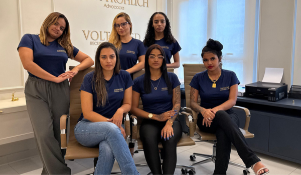 Seis integrantes da equipe Voltacred, de camiseta azul com o logotipo, reunidas na sala de atendimento da sede.